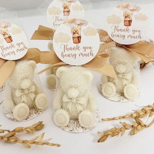 May include: White teddy bear favors wrapped in sheer fabric with a gold ribbon and a round tag that says "Thank you beary much" with a hot air balloon illustration.