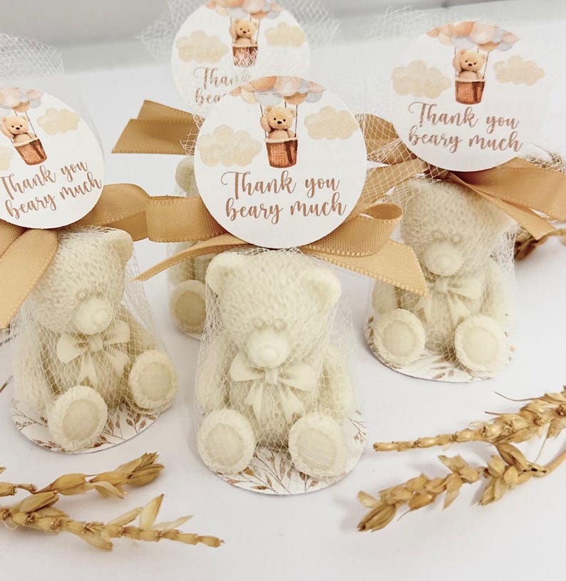 May include: White teddy bear favors wrapped in sheer fabric with a gold ribbon and a round tag that says "Thank you beary much" with a hot air balloon illustration.