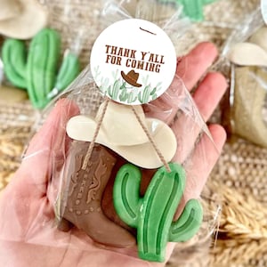 May include: A small gift bag filled with a brown cowboy boot, a green cactus, and a white cowboy hat. The bag has a round tag attached that says "Thank y'all for coming".