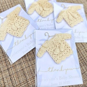 May include: Thank you cards with a crocheted baby sweater attached. The cards are white with blue and gold accents. The text on the cards reads "Thank you Javiersito's Baby Shower June 25 2022".
