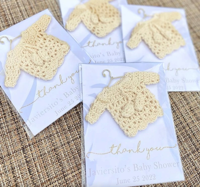 May include: Thank you cards with a crocheted baby sweater attached. The cards are white with blue and gold accents. The text on the cards reads "Thank you Javiersito's Baby Shower June 25 2022".