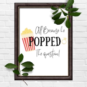 May include: A white sign with black text that reads "All Because he POPPED the question!" with a cartoon image of a red and white striped popcorn bucket with yellow popcorn. A gold ring with a diamond is next to the word "POPPED".