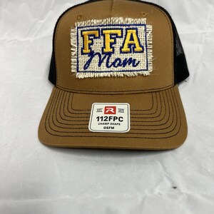 May include: A brown trucker hat with a black mesh back. The front features a patch with "FFA Mom" in yellow and blue lettering. A small tag at the bottom reads "112FPC CHAMP SHAPE OSFM".