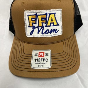 May include: A brown trucker hat with a black mesh back. The front features a patch with "FFA Mom" in yellow and blue lettering. The hat also has a tag that reads "112FPC CHAMP SHAPE OSFM".