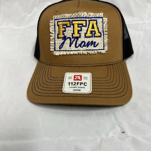 May include: A brown trucker hat with a black mesh back. The front features a patch with "FFA Mom" in yellow and blue lettering. A small tag at the bottom reads "112FPC CHAMP SHAPE OSFM".