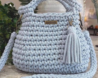 May include: A light blue crochet handbag featuring a thick braided handle and a matching detachable shoulder strap. The bag has a textured stitch pattern, a small rectangular metal tag labeled hand made, and a decorative tassel attached to a brass ring.