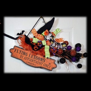 May include: A Halloween-themed witch's broom decoration with a black witch hat, orange and green ribbons, and a sign that reads "Flying Lessons by Appointment Only." The broom features orange and purple accents and black decorative balls.