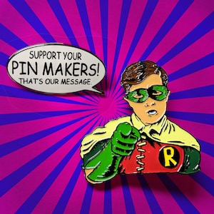 May include: A red, yellow, and green enamel pin featuring a cartoon character with a green mask and a red cape. The character is pointing with one hand and the other hand is holding a red and yellow 'R' on a yellow background. The pin has the text 'Support Your Pin Makers! That's Our Message.'
