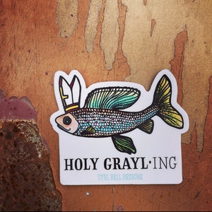 Puede incluir: Una pegatina con una ilustración colorida de un pez con una corona. El texto "HOLY GRAYL:ING" y "STEL BELL DESIGNS" está impreso debajo del pez.