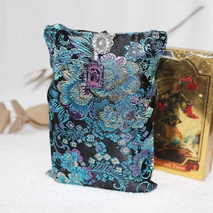May include: A black satin tarot card bag with teal and purple floral embroidery. The bag has a silver flower clasp and a small purple charm. A gold tarot card box is visible in the background.