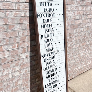 May include: A wooden sign with a white background and black text. The sign lists the NATO phonetic alphabet, starting with Alpha and ending with Zulu.