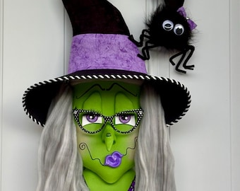 Puede incluir: Una decoración de pared que muestra a una bruja de cara verde con cabello gris, usando un sombrero negro y morado con una pequeña araña negra. La bruja tiene labios morados, gafas con montura negra y un corpiño a rayas blancas y negras.