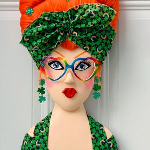 May include: A decorative wall hanging featuring a stylized face with orange hair, a green bow, and rainbow-colored heart-shaped glasses. The face has blue eyes, red lips, and shamrock earrings. The top is green with a shamrock pattern.