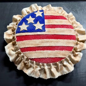 May include: A round, patriotic decoration featuring the American flag design. The center has red and tan stripes with a blue field and white stars. The edge is trimmed with a ruffled, tan fabric.