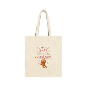 May include: Cream-colored tote bag with the text "Just a girl who loves her cavapoo" in pink and a cartoon image of a brown cavapoo dog. The bag has two handles and is made of a canvas-like material.