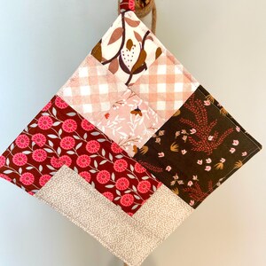 May include: A quilted, diamond-shaped pot holder with a wooden ring for hanging. The patchwork design features various floral and geometric patterns in shades of pink, brown, and cream. The pot holder is suspended by a natural rope.