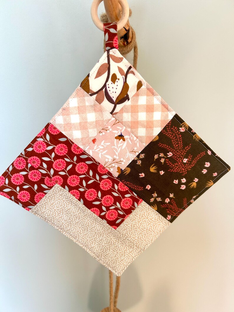 May include: A quilted, diamond-shaped pot holder with a wooden ring for hanging. The patchwork design features various floral and geometric patterns in shades of pink, brown, and cream. The pot holder is suspended by a natural rope.