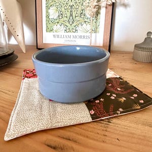 May include: A light blue ceramic bowl sits on a patchwork trivet with floral and dotted patterns. The trivet is made of fabric and has a pink border. A framed print with the text "WILLIAM MORRIS LONDON" is in the background.