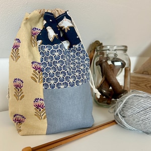 Peut inclure: Un sac à cordon avec un design en patchwork composé d'un imprimé floral beige, d'un motif géométrique bleu foncé et d'un panneau bleu clair uni. Le sac est fermé par des cordons blancs et présente un motif d'oiseau sur la partie bleue.