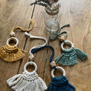 Può includere: Quattro giocattoli ad anello in legno fatti a mano con frange in macramé di colore giallo, bianco, verde e blu. I giocattoli sono appesi a una corda e sono esposti su una superficie di legno.
