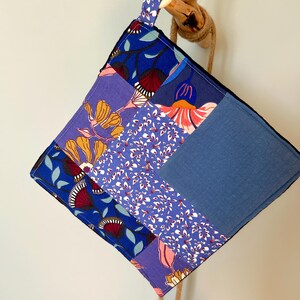 May include: A square, patchwork hot pad with a wooden ring for hanging. The fabric features a variety of blue floral patterns and solid blue sections. A natural rope is attached to the wooden ring.