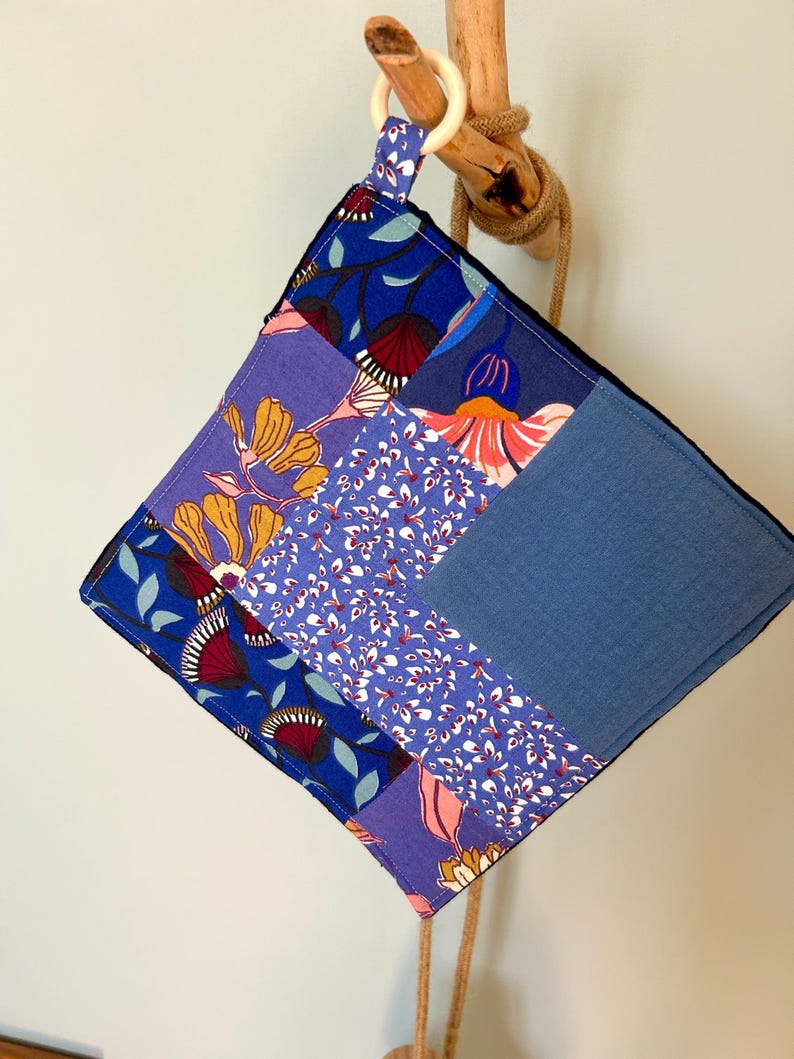 May include: A square, patchwork hot pad with a wooden ring for hanging. The fabric features a variety of blue floral patterns and solid blue sections. A natural rope is attached to the wooden ring.