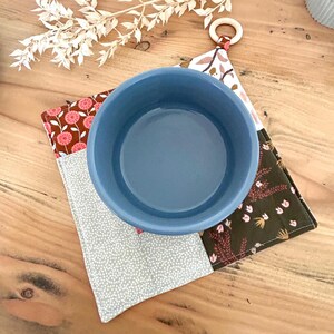 May include: A blue ceramic bowl sits atop a patchwork pot holder with floral and dotted patterns. The pot holder has a wooden ring for hanging. The color palette includes shades of blue, brown, pink, and gray, creating a rustic, handmade aesthetic.