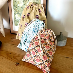 Op de afbeelding: Vier stoffen trekkoordzakken met bloempatronen. De tassen hebben een beige, witte en oranje kleurenpalet. De tassen hebben witte trekkoorden. De tassen zijn gerangschikt op een houten oppervlak.