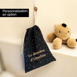 Könnte beinhalten: Ein schwarzer Beutel mit Kordelzug, weißem Sternenmuster und dem goldenen Schriftzug "Le doudou d'Opaline", der an einem Holzhaken neben einem beigen Plüschtier hängt. Ein schwarzes Feld mit weißem Text zeigt "Personnalisation en option".