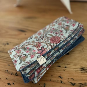Può includere: Un articolo in tessuto rettangolare piegato con uno sfondo verde menta chiaro e un denso motivo floreale nei toni del rosa, rosso e verde tenue. I bordi sono rifiniti con cuciture blu scuro e una piccola etichetta rettangolare bianca.