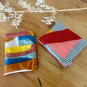Peut inclure: Deux pochettes rectangulaires en tissu avec des motifs en patchwork. L'une présente des bandes horizontales blanches, jaunes, bleu clair, bleu foncé et marron. L'autre comporte des panneaux diagonaux à rayures bleu marine, rouge et rose.