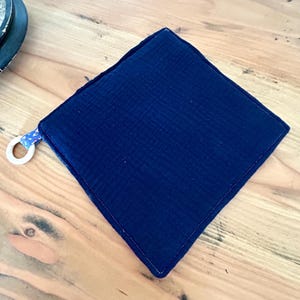 May include: A square, navy blue pot holder with a textured surface and a white wooden ring attached to a corner. The pot holder is made of a soft, quilted fabric and has a dark blue trim.