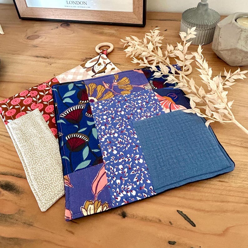 May include: A collection of colorful, patchwork-style pot holders. The designs include floral patterns in shades of blue, pink, and yellow, with a solid blue section. One pot holder has a wooden ring for hanging.