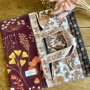 Peut inclure: Un sac fourre-tout en tissu avec un panneau bordeaux orné d'illustrations botaniques jaunes et orange, un panneau central à motif d'arbres blancs et marron, et une bande latérale bleu foncé à fleurs orange. Une étiquette indique Atelier du Chat Qui Pique.