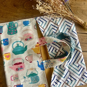Può includere: Una borsa in tessuto con un lato color crema caratterizzato da un motivo colorato di varie teiere e tazze, e un lato con un motivo geometrico a linee blu e marroni. Una piccola etichetta bianca riporta la scritta ATELIER DU CHAT QUI PIQUE.