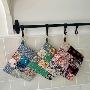 Peut inclure: Trois maniques carrées matelassées suspendues à une barre en métal noir. Chaque manique présente un motif patchwork avec divers imprimés floraux et géométriques dans des tons de bleu, vert, rose et noir, avec un anneau en bois clair.