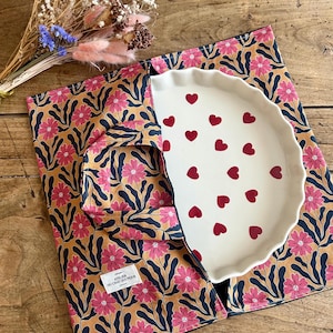 Pode incluir: Um suporte de tecido com fundo amarelo mostarda, flores cor-de-rosa e folhas azul-escuras, com uma etiqueta branca que diz ATELIER DU CHAT QUI PIQUE. Dentro, há uma assadeira de cerâmica branca com bordas onduladas e corações vermelhos.