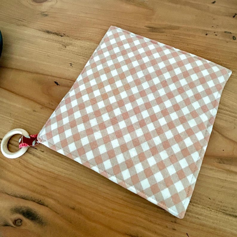 May include: A square, handmade baby teether with a peach and white gingham pattern. The teether has a wooden ring attached with a red and white patterned ribbon. The teether is laying on a wooden surface.
