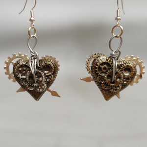 May include: A pair of gold-toned earrings featuring a heart-shaped charm with intricate gear details and a small arrow accent.