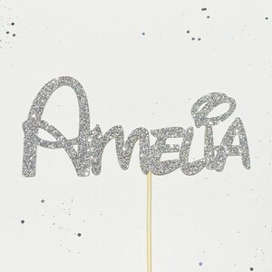 Puede incluir: Topper de pastel de purpurina plateada con el nombre Amelia en una fuente caprichosa.
