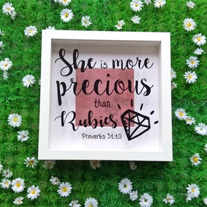 May include: White framed box with a pink background and black text that reads "She is more precious than rubies Proverbs 31:10". A black diamond is drawn next to the text.