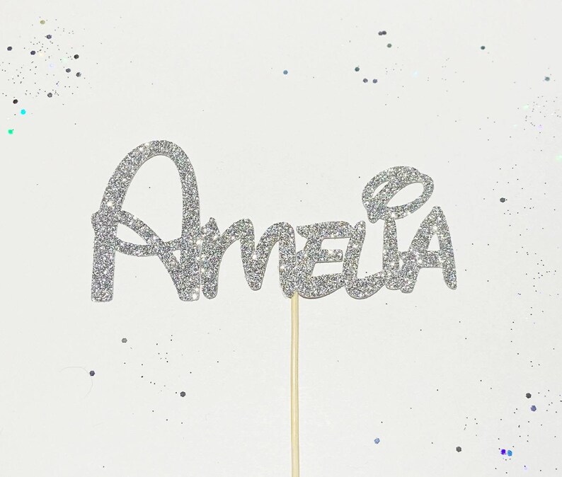 Puede incluir: Topper de pastel de purpurina plateada con el nombre Amelia en una fuente caprichosa.