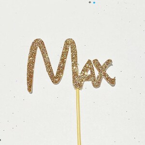 Puede incluir: Topper de pastel de purpurina dorada con el nombre "Max" escrito en cursiva.