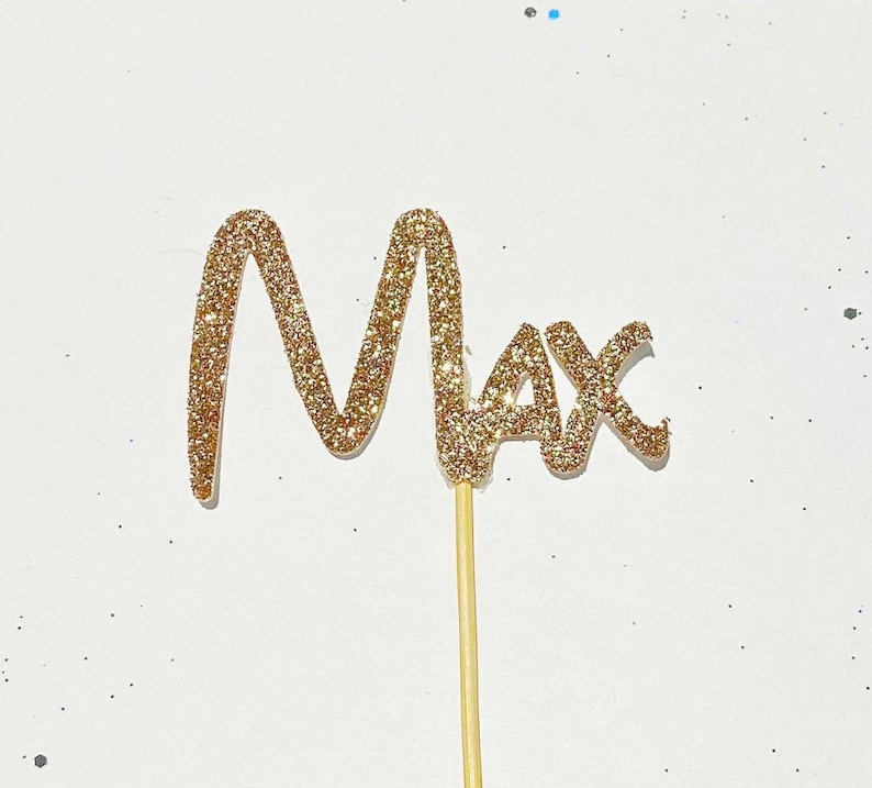 Puede incluir: Topper de pastel de purpurina dorada con el nombre "Max" escrito en cursiva.