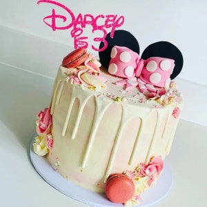 Puede incluir: Un pastel blanco decorado con detalles rosas y dorados. El pastel tiene un glaseado de chocolate blanco, chispas y macarons. Est&aacute; coronado con un lazo rosa y orejas negras, que recuerdan a Minnie Mouse. El adorno del pastel dice "Darcey 3".