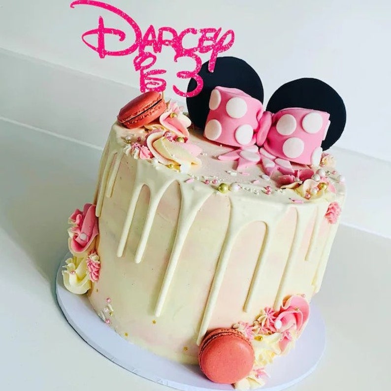 Puede incluir: Un pastel blanco decorado con detalles rosas y dorados. El pastel tiene un glaseado de chocolate blanco, chispas y macarons. Est&aacute; coronado con un lazo rosa y orejas negras, que recuerdan a Minnie Mouse. El adorno del pastel dice "Darcey 3".