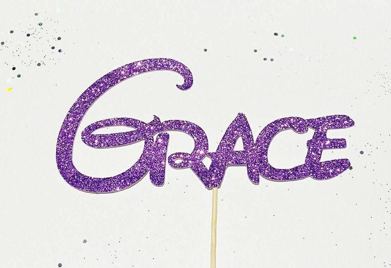 Puede incluir: Topper de pastel de purpurina morada con la palabra "GRACE" en una fuente caprichosa.