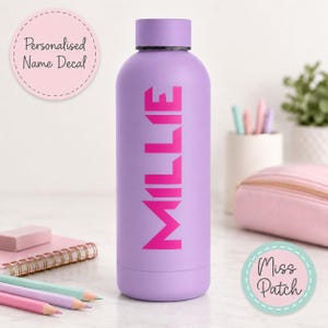 May include: A light purple water bottle with a matching screw-top lid. The name MILLIE is printed vertically in large, bold, hot pink capital letters on the front of the bottle. The bottle has a smooth, matte finish and a cylindrical shape.