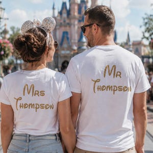 K&ouml;nnte beinhalten: Zwei wei&szlig;e kurz&auml;rmelige T-Shirts mit goldener Schrift auf der R&uuml;ckseite. Ein Shirt zeigt den Text Mrs Thompson und das andere den Text Mr Thompson. Die T-Shirts werden von zwei Personen getragen, die nebeneinander in einem &ouml;ffentlichen Bereich stehen.