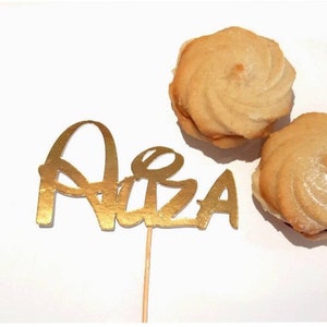 Puede incluir: Topper de pastel de purpurina dorada con el nombre "Alba" en un palo de madera. Dos galletas de mantequilla con glaseado blanco est&aacute;n en el fondo.